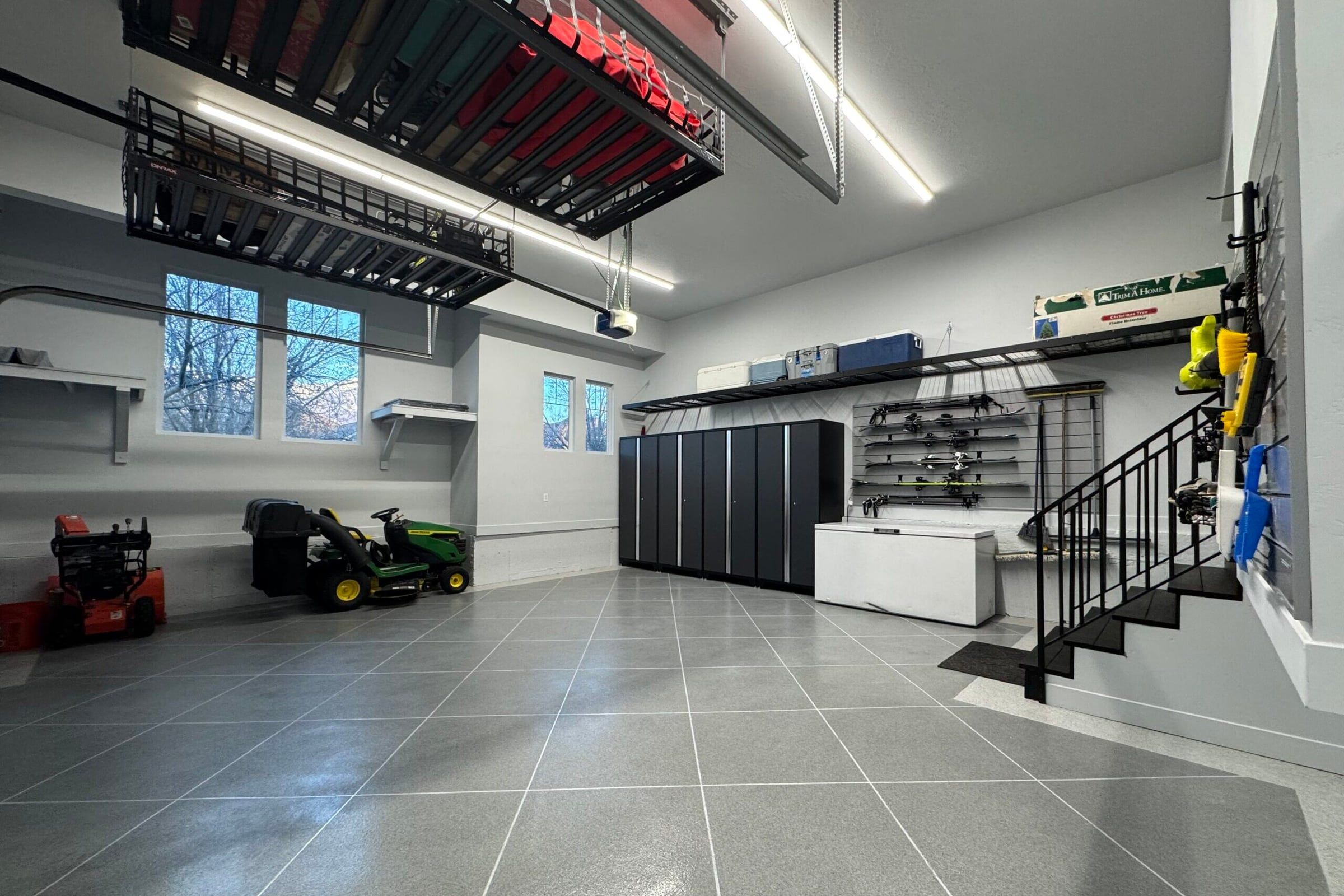 Large garage with grey tile floor, black cabinets, overhead racks and a stair rail