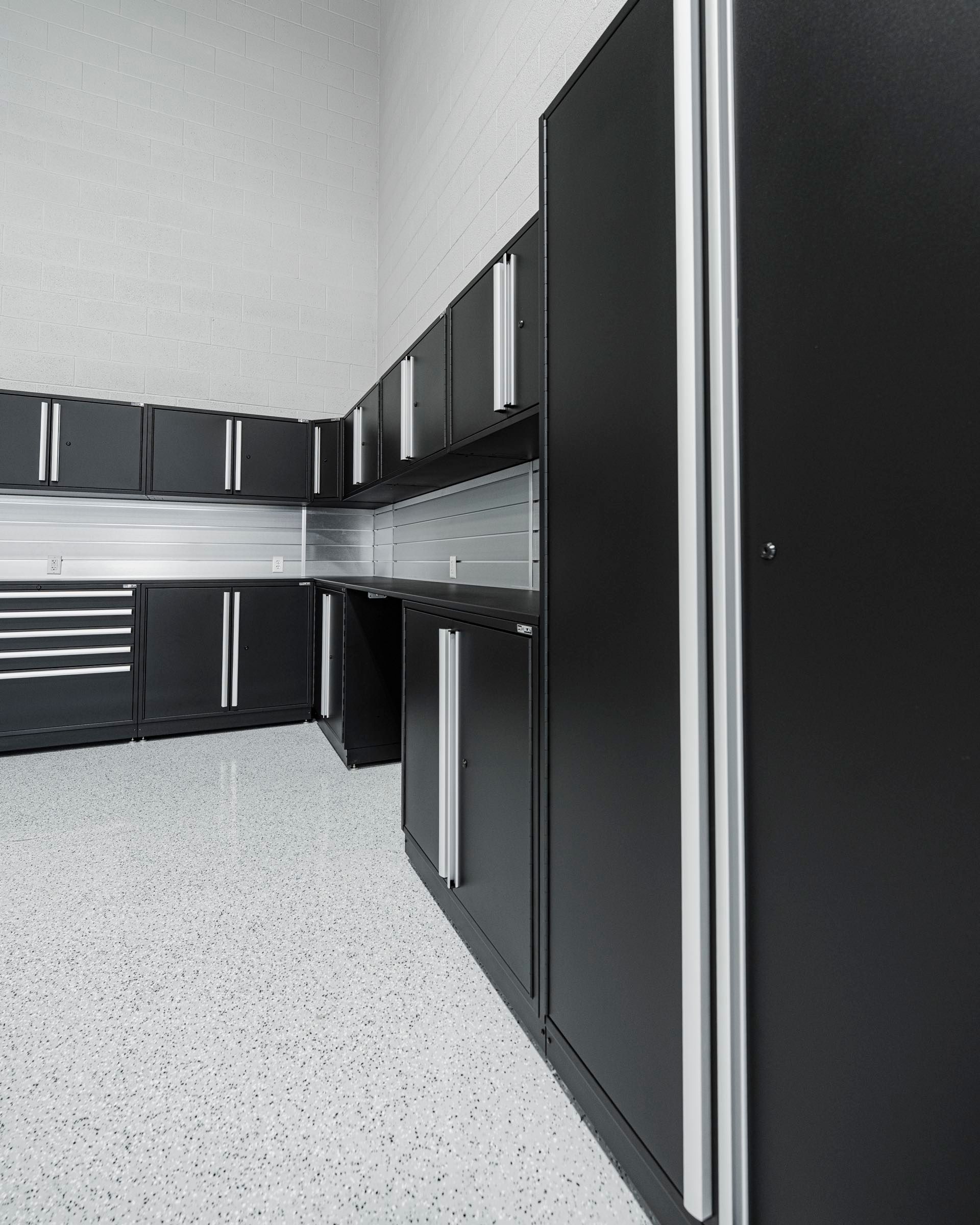 Corner run of black steel garage cabinets with silver handles, a stainless backsplash and tall locker cabinets on a flake floor