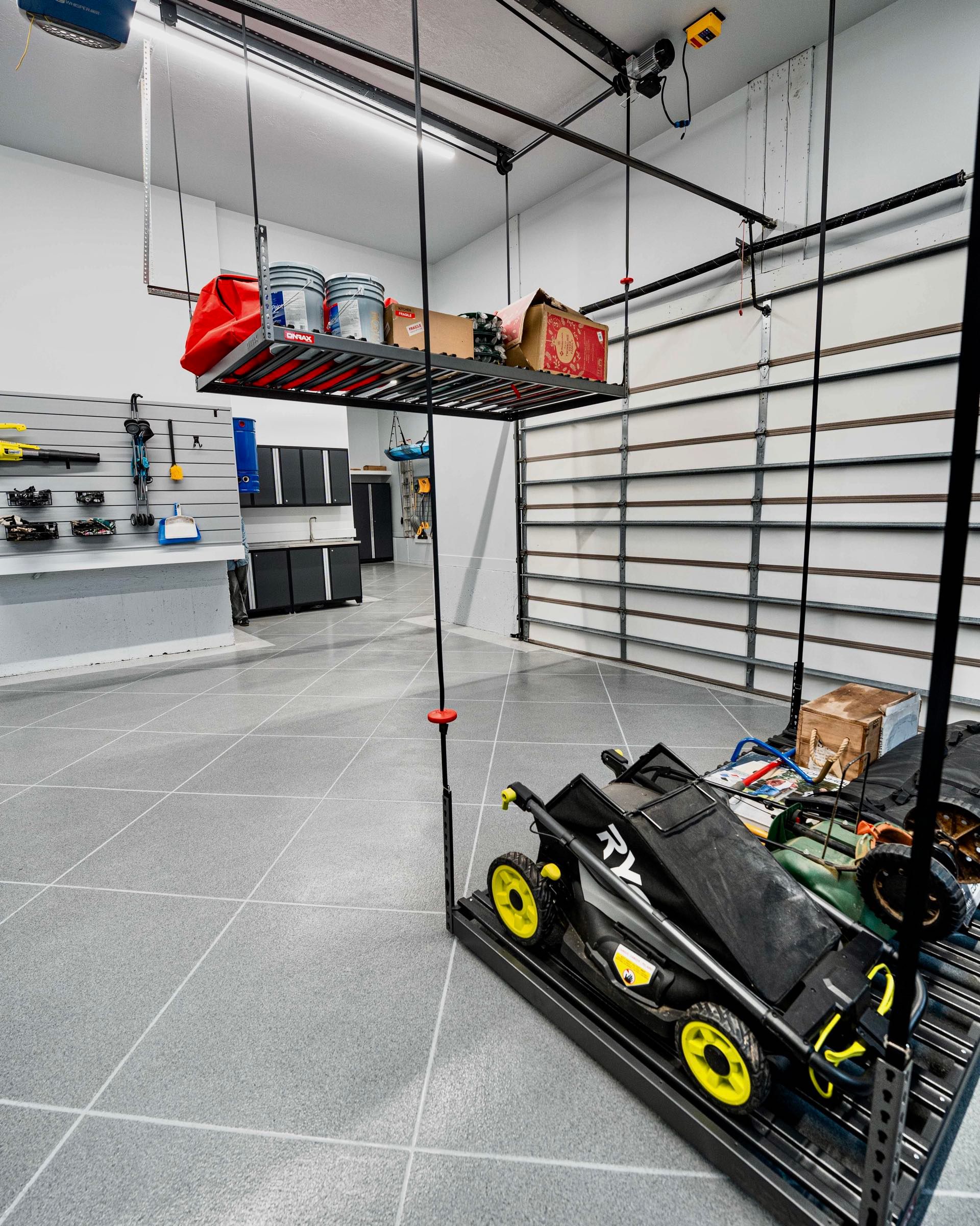 Cable-hung storage platforms in a garage, one holding buckets and boxes up near the ceiling and one holding a lawn mower, over a grey tile floor