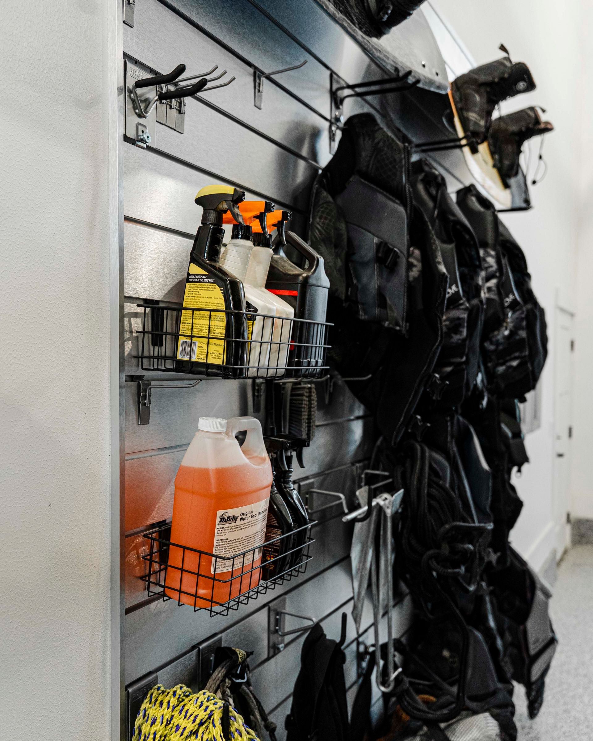 Metal slatwall with wire baskets of cleaning supplies, hooks, life vests and a wakeboard hung neatly on a garage wall