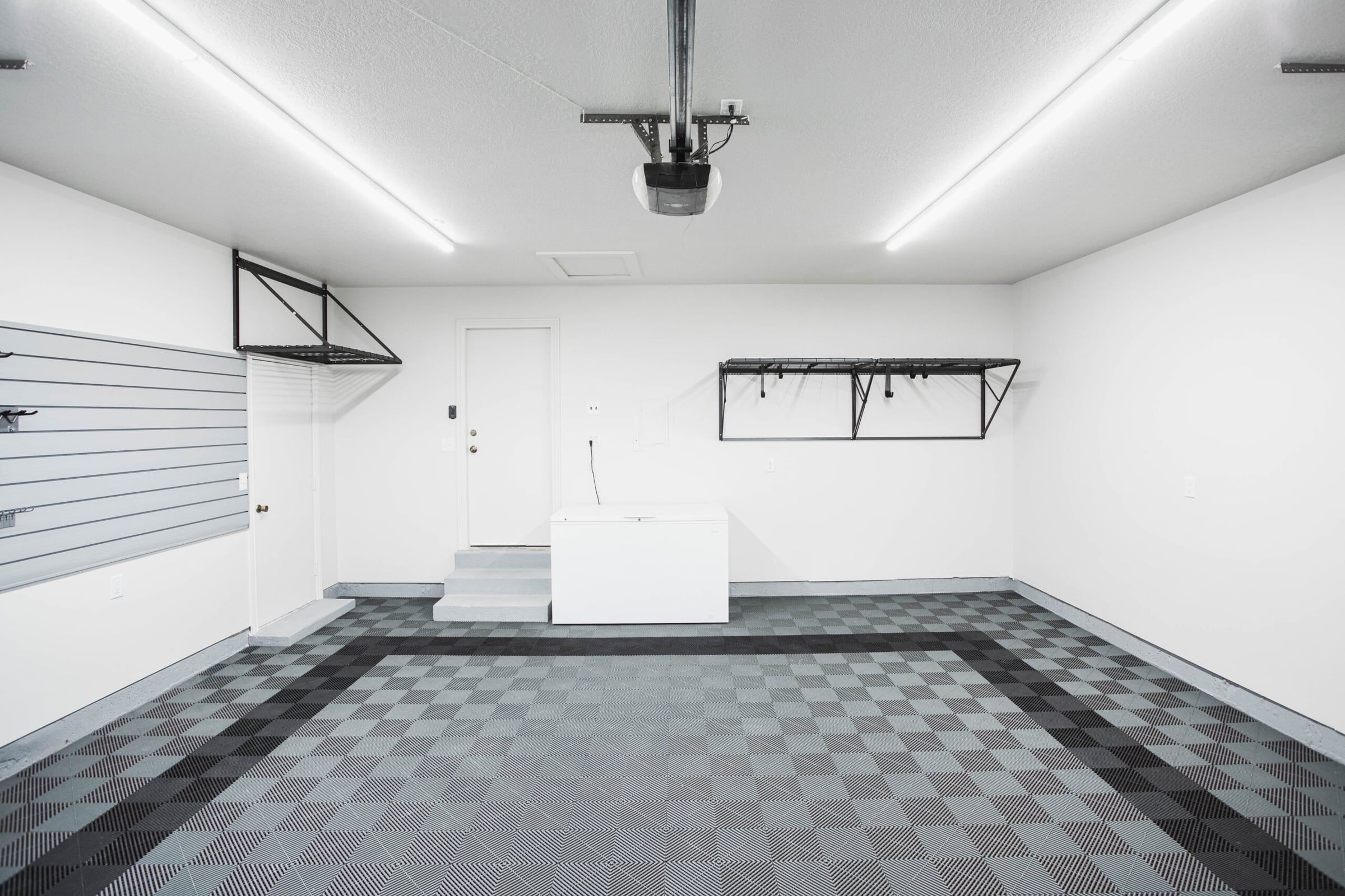After: the same garage with white painted walls, a grey checkered tile floor, wall shelving and LED lights